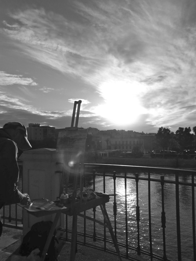A black and white shot of the artist on location in the city of Taranto in Puglia, Italy. The picture has been taken directly into the sunlight and the artist (Rupert Denyer) is seen as a silhouette. He is making a painting of the old city in Taranto.