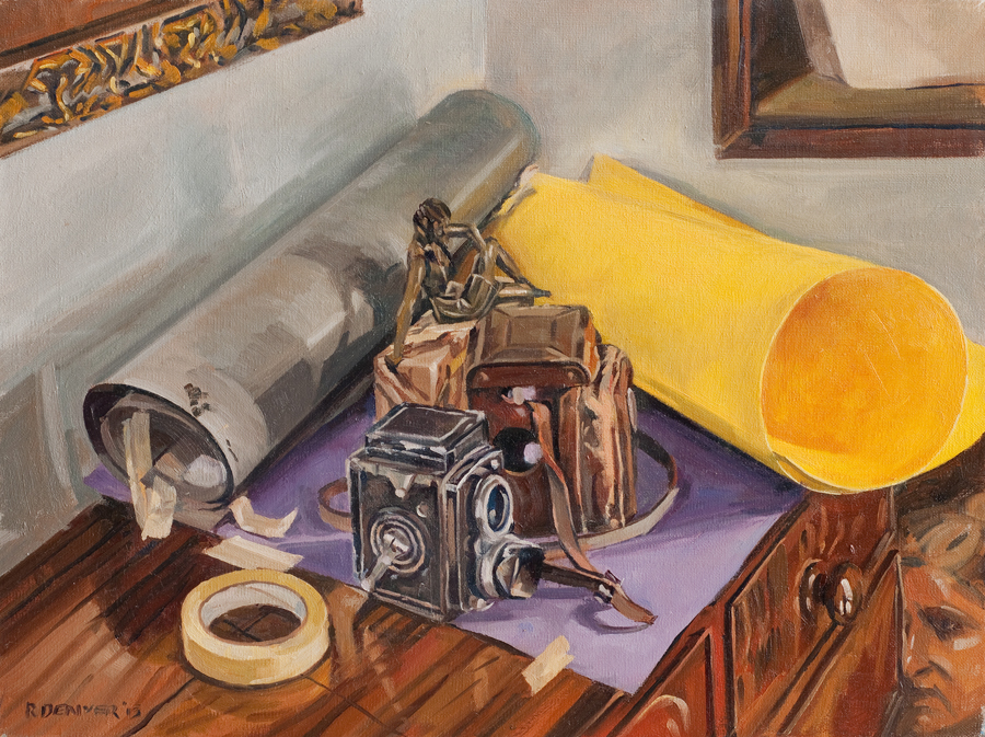 A still life sits on top of a brown wooden chest of drawers. there are two rolls of paper in the background, one yellow and the other grey. A small figurine, roliflex camera and case sit on top of a sheet of purple paper. There is a roll of masking tape to the side. The painting has been made in oil paint and is very well observed.